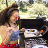 A person in a denim top and red pants spins vinyl at a DJ booth