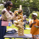 A parent and child in sunglasses play a game of jenga outdoors on a sunny day