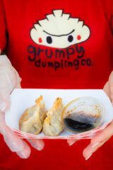 A person wearing a red shirt labeled "Grumpy Dumpling Co." offers a paper bowl of fried dumplings and sauce