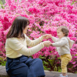 a woman is sitting on a stone wall in front of hot pink azaleas, holding hands with a little baby with yellow pants and hoodie