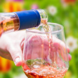 A person pours pink wine into a glass while surrounded by red and yellow flowers