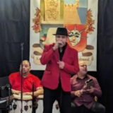 Three people in red shirts and jackets perform music on a stage