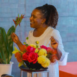 A person with long dark hair, wearing a white dress, creates a floral centerpiece with red and yellow flowers