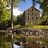 An old mill building made from stone with ten windows stands on a rocky river bank with a single, thin tree in the foreground and others in the background.