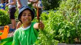 Young boy in blue hat and green shirt holding up a carrot with soil from the ground.