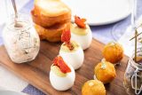 close up of different nosh snacks of fried round balls, hard boiled eggs, toast, and a dip in a mason jar on blue and white checkered table cloth, with a glass of rose wine