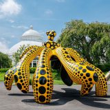 A massive pumpkin sculpture stylized as though it were dancing. Its body is orange with black polka dots, while its slightly twisted stem is black with orange polka dots. Its underside is solid black.