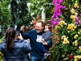 man holding child taking a photo on his phone while a woman is also taking a photo of the group in front of brign pink and yellow orchids