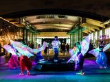 Performers with light up wings dancing around a DJ in front of the Visitor Center at night