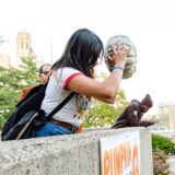 A person in a white shirt, with long dark hair, chucks a pale pumpkin from a rooftop
