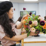 a person in a black barrette styling a bouquet of pink, orange, and dark purple roses in a classroom