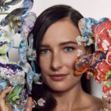 A person with long brown hair poses for a photo while surrounded by blue and red flowers