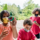 A child with medium-length brown hair wearing a mask and an orange shirt holding up a small green ball with beads on it resembling a face. A shorter child with short curly hair and a grey-and-white mask looks downward beside her, while another child with medium-length brown curly hair wearing a red shirt, black pants, and a black mask looks at some sort of green material in their hand.