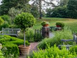 Pathway leading into Garden area with a short, red brick wall, a brick pathway, green potted tree, green bushes and two wooden benches with a hill of green grass and trees in the distance.