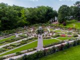 Aerial shot of Rose Garden with people walking on grey pathways, sections of green grass, rose bushes with small pink and white roses, green trees and blue skies.