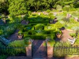 Aerial view of Garden area showing green potted trees, bushes around the perimeter, wooden benches in each corner, and bushes cut into geometric shapes in the center.