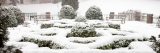 Snow covered bushes, benches, trees, hill and small brick pillars.