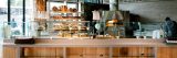 A cafe-style counter with sandwiches and bread behind glass, with a pizza oven in the background.