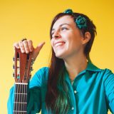 A headshot of Ali Dineen holding a guitar by the headstock and looking off to the side.