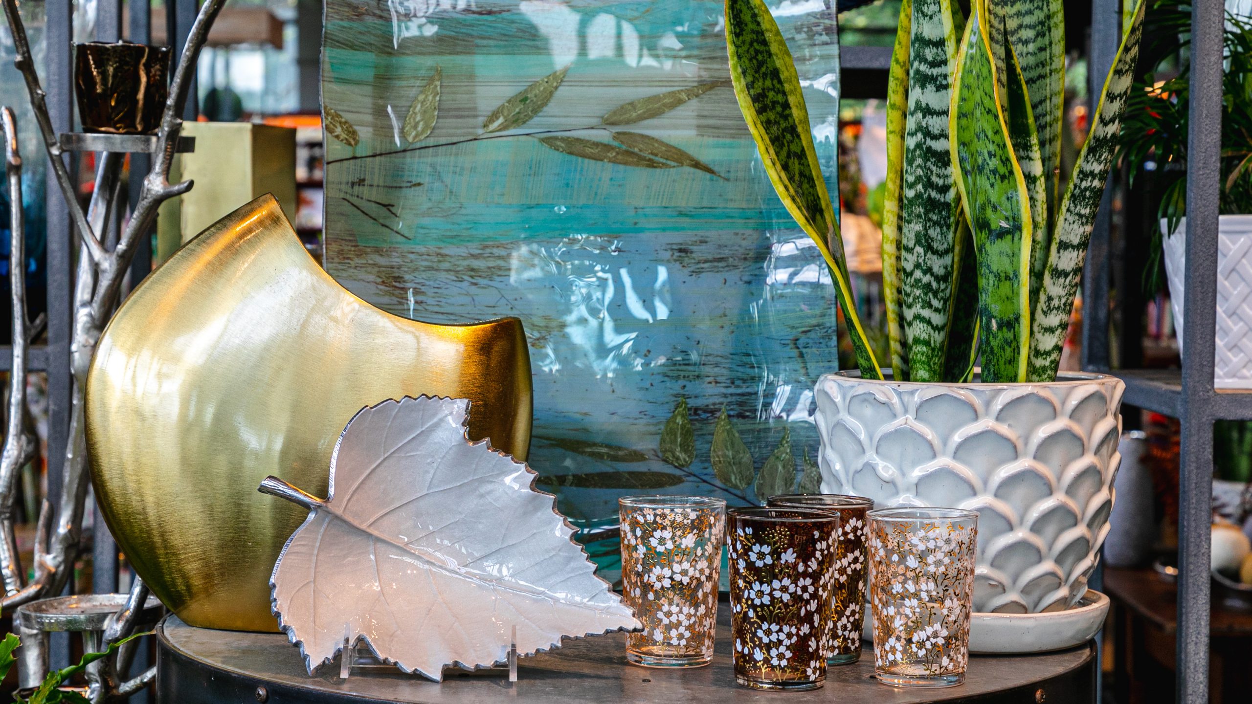 A variety of home decor, include floral glasses, a leaf-shaped dish, a riged white plant pot, a blue and gray serving tray, and a rounded, crescent-esque gold decorative sculpture