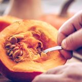 A person uses a spoon to scoop seeds out of the interior of an orange pumpkin