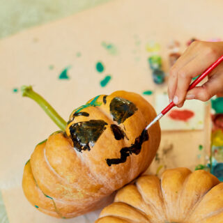 A person paints eyes and a mouth on a small orange pumpkin with a green stem