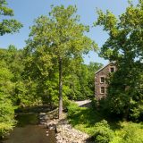 A stone mill beside a river with tall, lush trees growing around it.