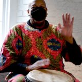 Image of african american man in bright red and gold sweater wearing a face mask holding one hand up while the other is on a large hand drum.
