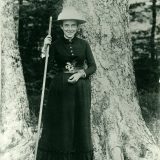 Black and white image of Elizabeth Britton standing outside next to a large tree with a triangular hat on and holding a large stick.