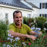 Headshot of Christopher Spitzmiller wearing an olive green shirt standing in front of purple flowers and greens with a white house behind him.