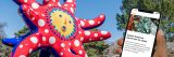 An image of a hand holding a phone in front of a tentacled red and blue statue with a yellow sun-like face and white polka dots.
