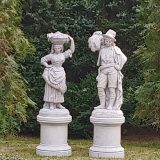 Female and male antique stone sculptures standing on pedestals, where the woman is holding part of her skirt with one hand and a basket on her head with the other, and the man looks like he is holding a sack. They are placed in a grassy lawn with high green trees all around.