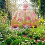 A large pumpkin sculpture covered in yellow mosaic tiles with red mosaic polka dots. Its stem is covered in red mosaic tiles with small yellow mosaic polka dots. The sculpture is nestled into a bed of low-lying flowers and plants, with small trees and vines rising up just behind it.