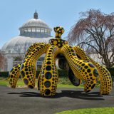 A massive pumpkin sculpture stylized as though it were dancing. Its body is orange with black polka dots, while its slightly twisted stem is black with orange polka dots. Its underside is solid black.