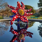 A large tentacled red and blue statue with a yellow sun-like face and white polka dots on its tentacles seems to hover above a reflecting pool, its image mirrored in the water. A mix of trees can be seen in the background.