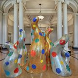 A group of tentacle-like sculptures covered in reflective golden or silver mosaic tiles, sporadically adorned with colorful polka dots.