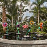 Five colorful floral sculptures of varying colors and styles rise up from a reflecting pool surrounded by various palm trees in a domed glasshouse.