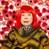 Yayoi Kusama, sporting bright red hair and a black shirt covered in light orange polka dots, sits in front of a white wall covered in red and white flowers.