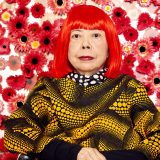 Yayoi Kusama, sporting bright red hair and a black shirt covered in light orange polka dots, sits in front of a white wall covered in red and white flowers.