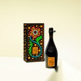 Black champagne bottle with orange lable for La Grande Dame 1917 resting on a white table cloth with the box for the bottle designed with a brightly colored polka dot Kusama flower behind it