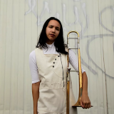 A young woman with dark hair standing in front of a white wall tagged with graffiti; she wears a matching white outfit, and holds a brass trombone