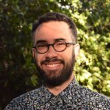 A bearded, light-skinned man in glasses smiles in front of a backdrop of bright green leaves