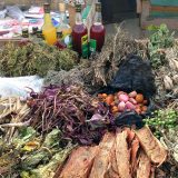 A table piled high with dried leaves, roots, bark, and fruits, with a selection of bottles filled with red and yellow beverages behind