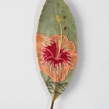 An ovate green leaf embroidered with the image of a tropical flower in yellow, pink, and red thread