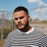 A bearded man with dark, close-cropped hair, wearing a horizontally striped black and white shirt, stands in front of a chainlink fence in a desolate urban scene