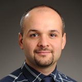 A man with upturned lips, a goatee, and a bald head poses for a headshot in front of a gray background, wearing a blue flannel shirt and dark tie