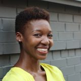 A woman with close-cropped dark hair wearing a yellow shirt poses in front of a gray brick wall for a photo, her lips upturned