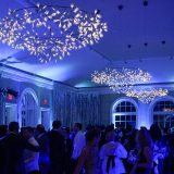 Guests in formal dress in a dim, blue-tinged room. Fairy lights dangle from metal-rimmed plain chandeliers on the ceiling.