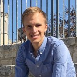 A light-haired man in a blue buttoned shirt poses for a photo in front of an iron gate framed by a blue sky