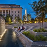 Two people sit at dusk on a series of stone steps leading down into a well-lit water feature, trees surrounding the space and the spike of the Washington Monument rising up in the distance